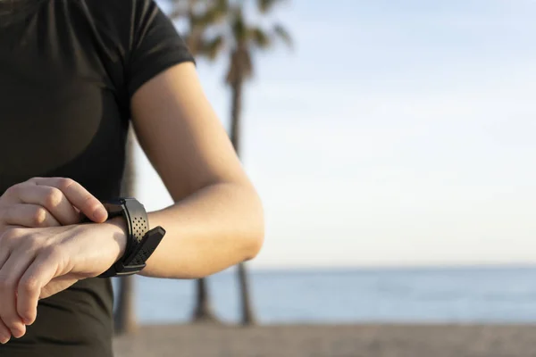 Young fitness woman using smart watch before or after running on beach background with palm trees.Sports wellness and athletics people lifestyle concept with copy space.