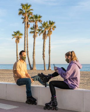 Tıbbi koruyucu maskeli patenci çift (Coronavirus 'a karşı, covid-19) gezinti güvertesinde oturuyor, deniz kenarında cep telefonu kullanıyor, palmiye ağaçları ve plaj geçmişi kullanıyor. Sağlıklı, fitness teknolojisi kavramı