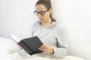 Genç beyaz kadın yatakta kitap okuyor.