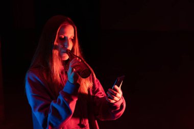 Bira içen genç bir kız ellerindeki akıllı telefona bakıyor. Stüdyo koyu arka planda beyaz bir kadının renkli ışıklı portresi. Fotokopi uzayı olan gece kulübü parti konsepti yaşam tarzı..
