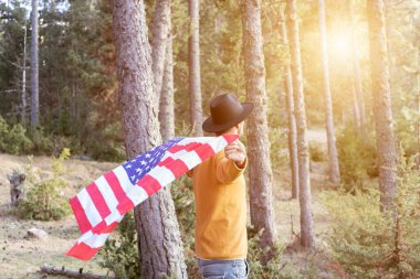 Siyah kovboy şapkalı bir adam gün batımında ormanda Amerikan bayrağı sallıyor. Güneş ışığı ve kopyalama alanı olan güzel bir orman manzarası. 4 Temmuz Bağımsızlık Günü kutlamaları konsepti..