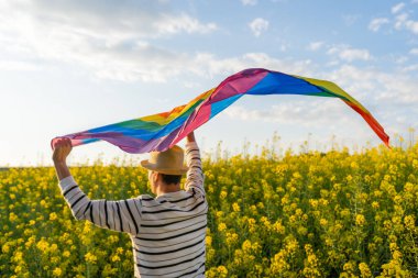 Gün batımında sarı çiçek tarlasında gökkuşağı bayrağı taşıyan bir adam. Elinde eşcinsel gururu bayrağı tutan bir erkek, LGBT kavramı. Güneşli bir gün kutlaması..