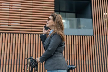 İş kadını portresi bisikletle işe giderken modern ofis arka planında akıllı telefonla konuşuyor..