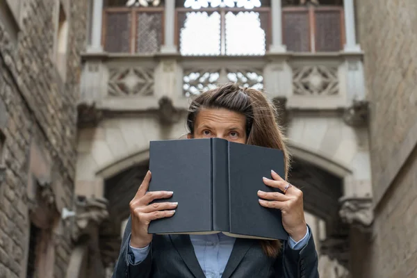 Young woman hides his face behind a book on ancient city building background.