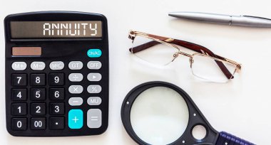 ANNUITY kelimesi bir hesap makinesi ve beyaz bir arkaplan üzerine yazılmıştır. Yakınlarda bir gözlük, bir kalem ve bir büyüteç var. İş konsepti