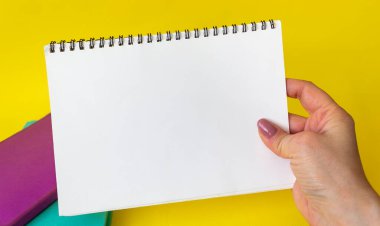 Kadının eli yazmak için boş bir not defteri tutuyor. Sarı arka planda renkli not defterleri var. Boşluğu kopyala