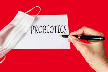 PROBIOTICS kelimesi bir kadın eli tarafından bir deftere yazılır. Kırmızı arka planda bir tıbbi maske yatar. Tıp konsepti