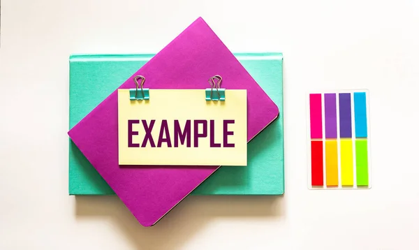 Example, the word is written on a sticker and a white background. The sticker lies on the example on colored notebooks.