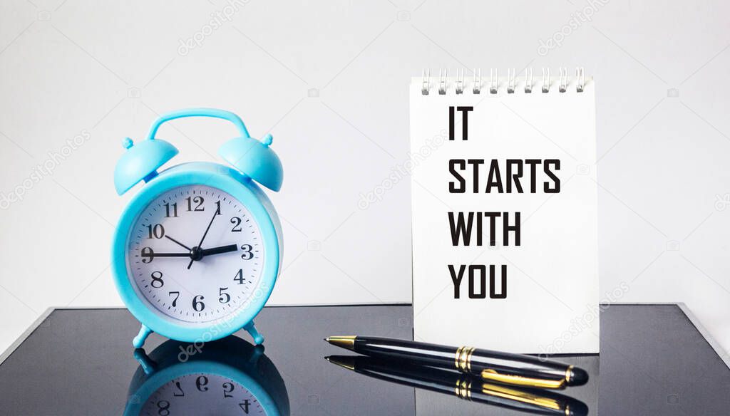 It all starts with you, text on a notebook, next to an alarm clock and a pen on a glass black background