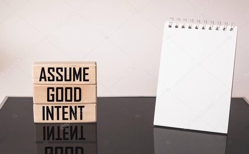 Assume good intent. Inspirational quote on wooden blocks with notepad to write on white and black background. Positivity concept