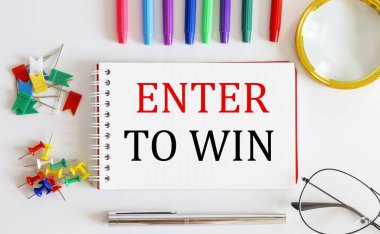 Beyaz masadaki ofis araçlarıyla ENTER To WIN sözcüğünü içeren defter.