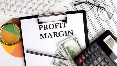 Klavye, dolar, hesap makinesi, malzeme ve beyaz arkaplan üzerinde analiz çizelgesi içeren ofis masasında PROFIT MARGIN metni.