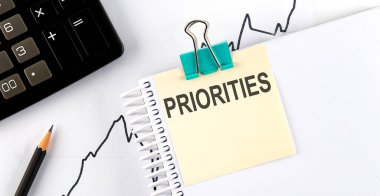 Beyaz arka planda, hesap makinesi ve ofis malzemelerinin yanında PRIORITIES yazılı not. İş konsepti.