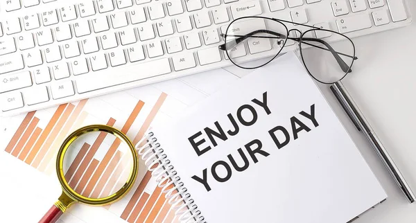 ENJOY YOUR DAY text written on notebook with keyboard, chart,and glasses