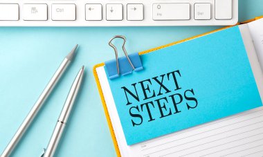 Mavi arkaplanda kalem ve klavye ile yapıştırılmış NEXT STEPS metni