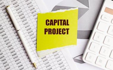 Sarı bir kağıda yazılmış sermaye projesi. Finansal belgeler, kalem ve hesap makinesi masanın üstünde.. 