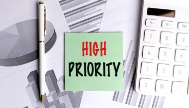 Hesap makinesi, kalem ve finansal tablolarla yeşil yapışkan nottaki yüksek öncelikli mesaj, iş görevlerindeki aciliyeti simgeliyor.