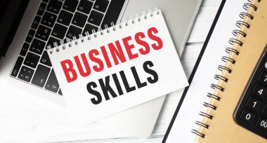 Klavyede keçeli kalemler ve tahta zemin üzerinde hesap makinesi olan BUSINESS SKILLS metni olan not defteri.