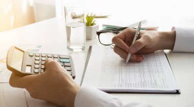 Ofis masasında hesap makinesiyle çalışan bir işadamı, bir kalem ve belge. Sayılar sayılıyor ve hesaplamalar yapılıyor.