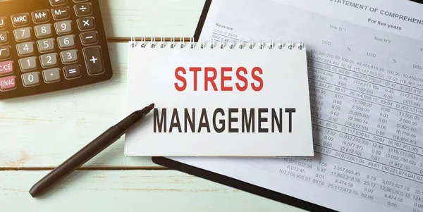 Stress management statement on paper notepad. Office desk with electronic devices, a document with a table below, a concept image for the blog title or a header image.