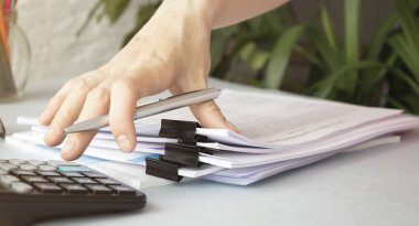 İş adamının elleri, kağıt yığınları halinde çalışmak için kalem tutar, iş raporları için bilgi arar ve modern bir ofisteki bir dizüstü bilgisayar masasında yığınla eksik belge elde edilir.