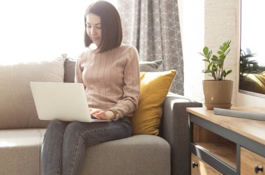 Gülümseyen genç kadın dizüstü bilgisayar kullanıyor, evdeki kanepede oturuyor.