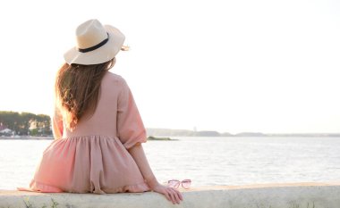 Arkadan görülen şapkalı kadın kayanın üzerinde oturuyor ve denize bakıyor.