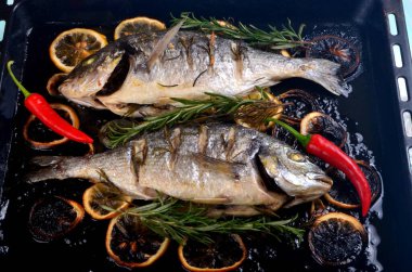 Lezzetli kavrulmuş iki dorado balığı, limon dilimleri, baharatlar ve siyah bir fırında taze biberiye.