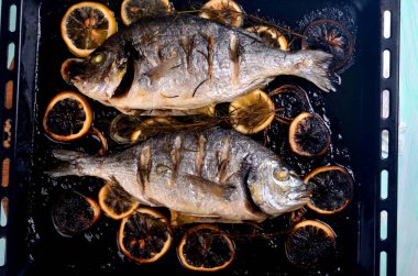 Lezzetli kavrulmuş iki dorado balığı, limon dilimleri, baharatlar ve siyah bir fırında taze biberiye.