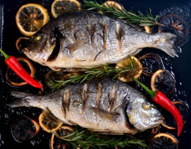 Lezzetli kavrulmuş iki dorado balığı, limon dilimleri, baharatlar ve siyah bir fırında taze biberiye.