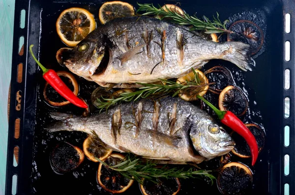 Lezzetli kavrulmuş iki dorado balığı, limon dilimleri, baharatlar ve siyah bir fırında taze biberiye.