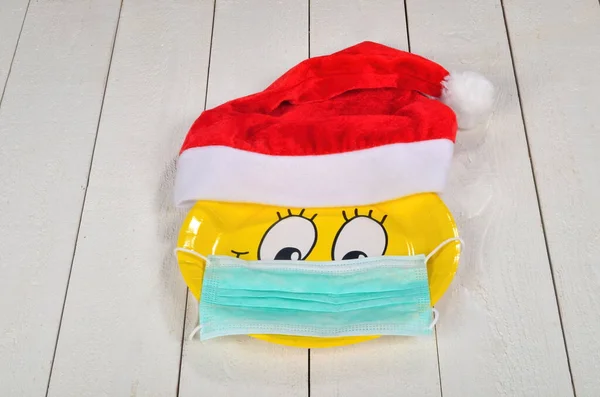 Happy smiley cartoon face on yellow paper plate and red Santa Claus hat, with face protection mask. White wooden background. Happy yellow emoticon smiling, korobavirus protection concept at Christmas