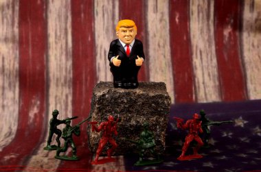 RIGA, LATVIA - HAZİRAN 07, 2021: Bir kayanın üzerinde duran küçük Donald Trump figürü, ayaklarının dibinde duran asker figürleri, klasik ABD bayrak alanı
