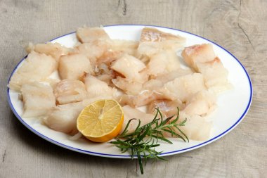 Limon ve biberiyeyle süslenmiş beyaz bir tabakta deniz balığı filetosu, tabakta beyaz çiğ balık, gri ahşap zemin.