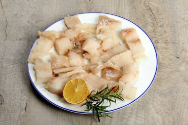 Limon ve biberiyeyle süslenmiş beyaz bir tabakta deniz balığı filetosu, tabakta beyaz çiğ balık, gri ahşap zemin.