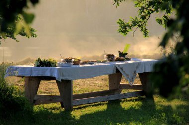 Gün batımında açık havada yaz sahnesi partisi masası. Ağaçların altında yemek tabağıyla eski ahşap bir masa. Letonya 'da yaz ortası kutlaması, Ligo festivali