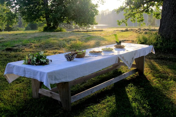 Outdoors summer scene party table on sunset. Old wooden table under trees with food plate. Midsummer celebrating in Latvia, Ligo festive