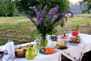 Yaz ortası yemek masası. Ağaçların altındaki bahçede içecekler ve yiyeceklerle dolu bir masa. Masanın üzerinde mavi çayır çiçekleri olan cam bir vazo var. Letonya LIGO Festivali