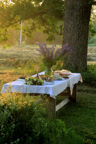 Midsummer food table.Table filled with drinks and food outside in the garden under the trees. on the table a glass vase with blue meadow flowers. Latvian LIGO festive