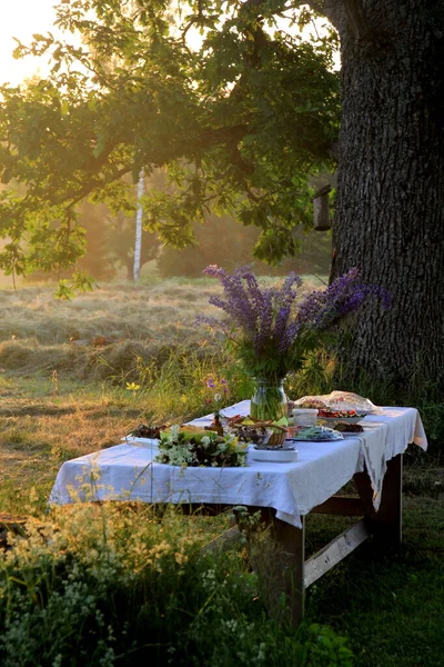 Yaz ortası yemek masası. Ağaçların altındaki bahçede içecekler ve yiyeceklerle dolu bir masa. Masanın üzerinde mavi çayır çiçekleri olan cam bir vazo var. Letonya LIGO Festivali