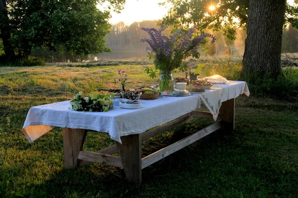 Yaz ortası yemek masası. Ağaçların altındaki bahçede içecekler ve yiyeceklerle dolu bir masa. Masanın üzerinde mavi çayır çiçekleri olan cam bir vazo var. Letonya LIGO Festivali