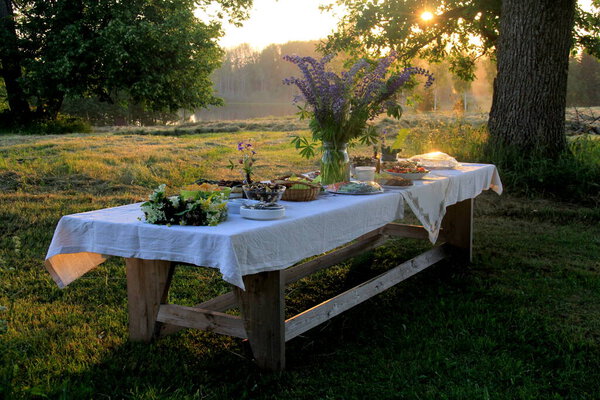 Стол с напитками и едой в саду под деревьями. на столе стеклянная ваза с голубыми лугами. Праздник Латвийского ЛИГО