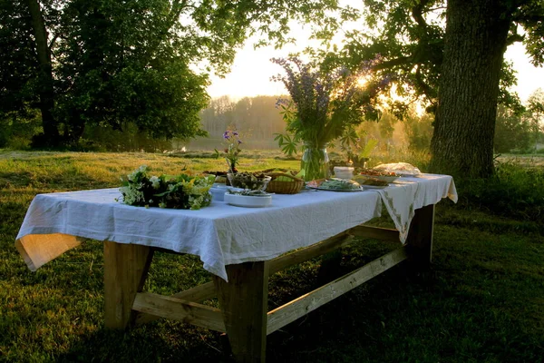 Yaz ortası yemek masası. Ağaçların altındaki bahçede içecekler ve yiyeceklerle dolu bir masa. Masanın üzerinde mavi çayır çiçekleri olan cam bir vazo var. Letonya LIGO Festivali