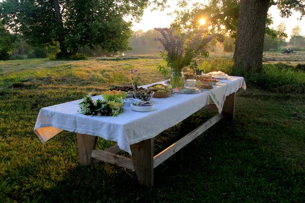 Yaz ortası yemek masası. Ağaçların altındaki bahçede içecekler ve yiyeceklerle dolu bir masa. Masanın üzerinde mavi çayır çiçekleri olan cam bir vazo var. Letonya LIGO Festivali