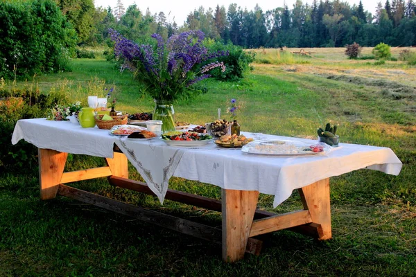 Yaz ortası yemek masası. Ağaçların altındaki bahçede içecekler ve yiyeceklerle dolu bir masa. Masanın üzerinde mavi çayır çiçekleri olan cam bir vazo var. Letonya LIGO Festivali