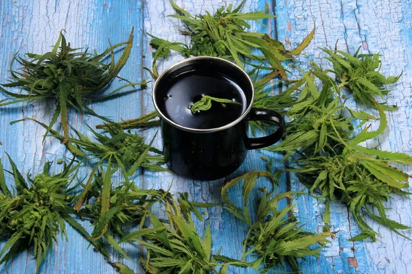 Marijuana herbal tea in a cups and green cannabis leaves around. Blue painted wooden board background. Hemp Tea. Cannabis herbal tea