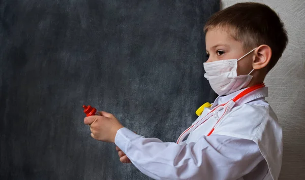 A charming little boy in a medical mask and white uniform holds a toy thermometer in his hand. Not sharp focus, dark background. Doctor game, future profession concept