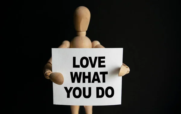 The wooden man holds a white sign with the text LOVE WHAT YOU DO in his hands. The content of the lettering has implications for business concept and marketing.