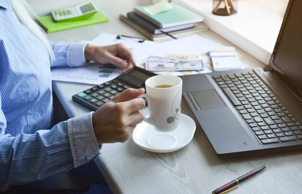 Female hands hold documents and business graphics on the background of a laptop. The girl works in the home office at the table.