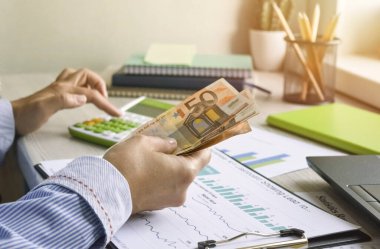 Kadın elleri, yaklaşın. Bir muhasebeci olan iş kadını para sayıyor ve elinde bir banknot tutuyor. Dizüstü bilgisayarlı ve grafikli çalışma masası.